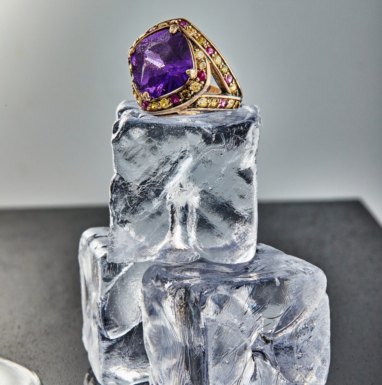 Yellow gold ring with amethyst on ice cubes on a gray background with reflection. Jewelry art and product sales.